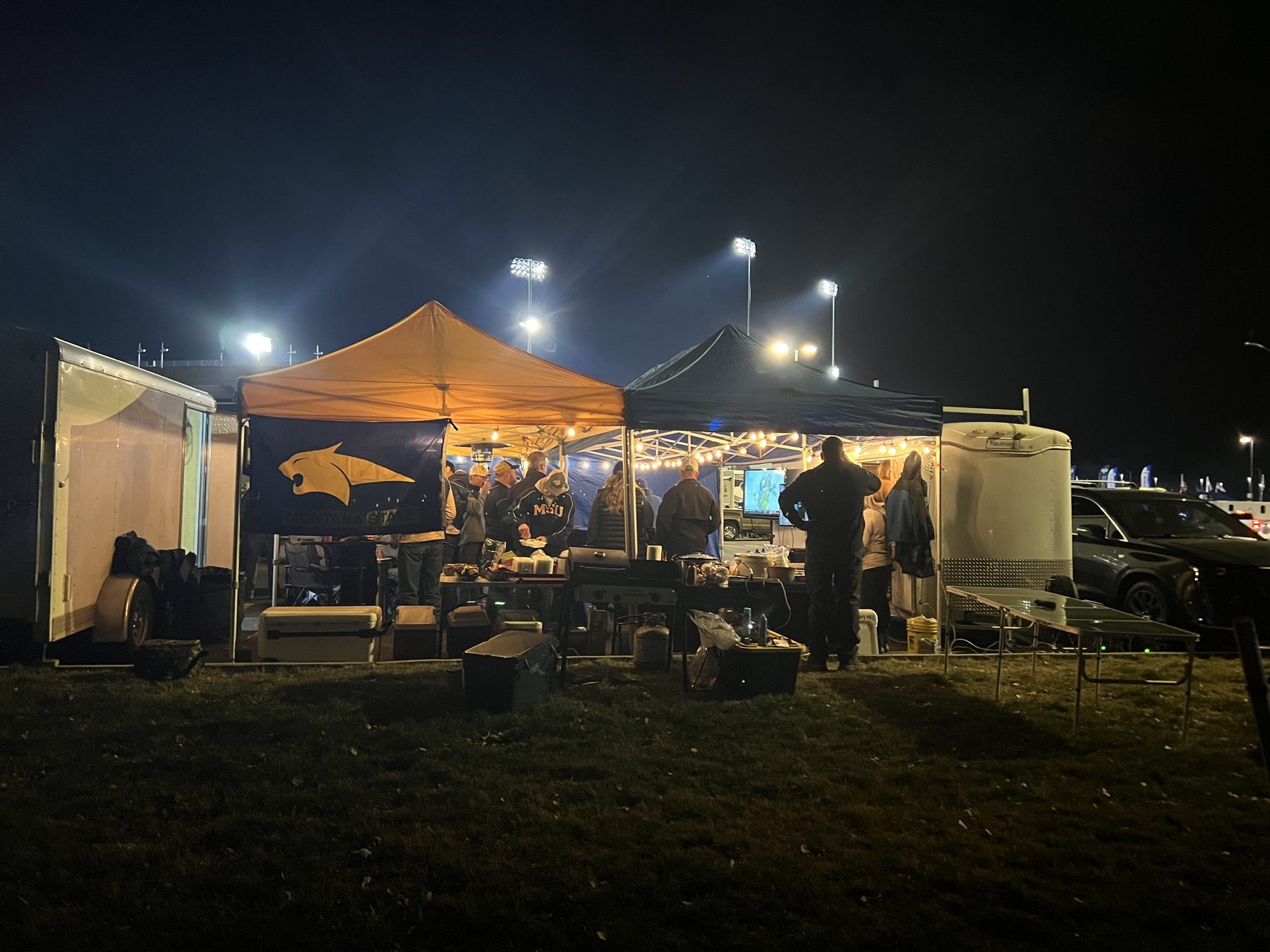 Bobcat fans tailgating at night under string lights and the Bobcat Stadium light towers in Bozeman